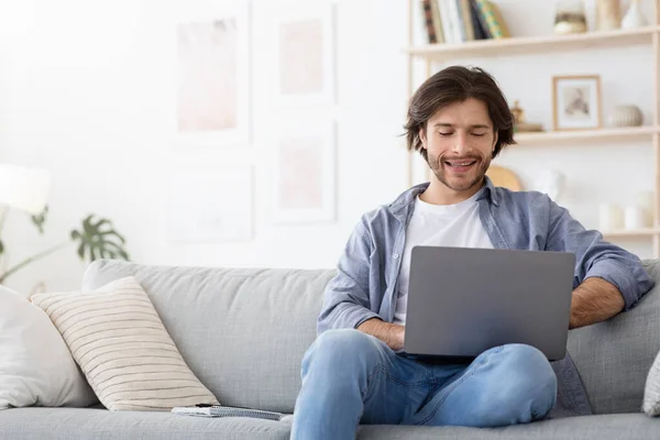 Веселый парень сидит дома на диване, используя ноутбук — стоковое фото