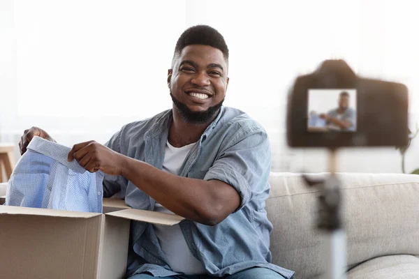 Ξεκαρδιστικό Vlog. Χαμογελώντας μαύρο άντρα ξεπακετάρισμα κουτί με νέα ρούχα στην κάμερα — Φωτογραφία Αρχείου