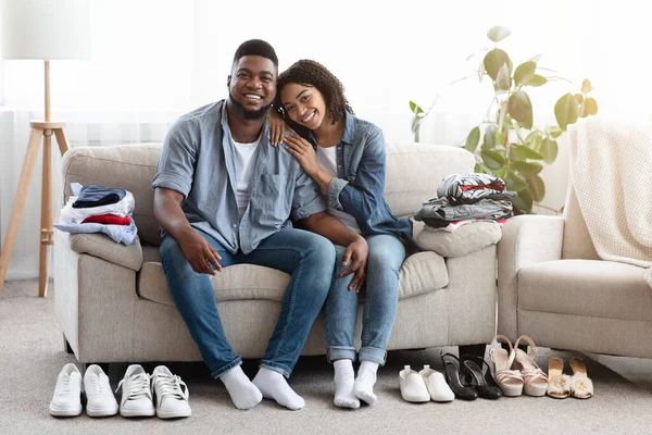 Felice coppia nera seduta sul divano con vestiti e scarpe piegati intorno — Foto Stock