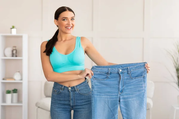 快乐瘦长的女人站在家里展示超大的旧牛仔裤 — 图库照片