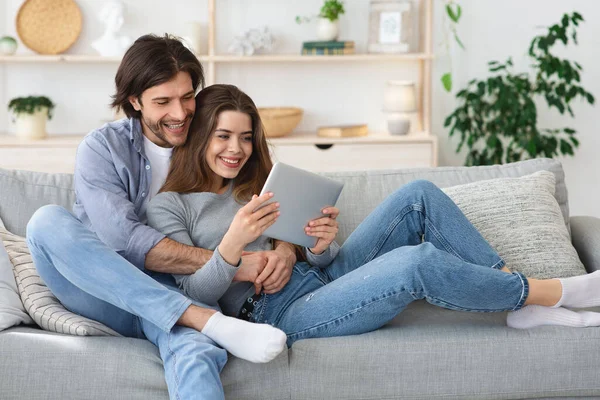 Alegre pareja abrazándose en el sofá en casa, utilizando la tableta digital —  Fotos de Stock