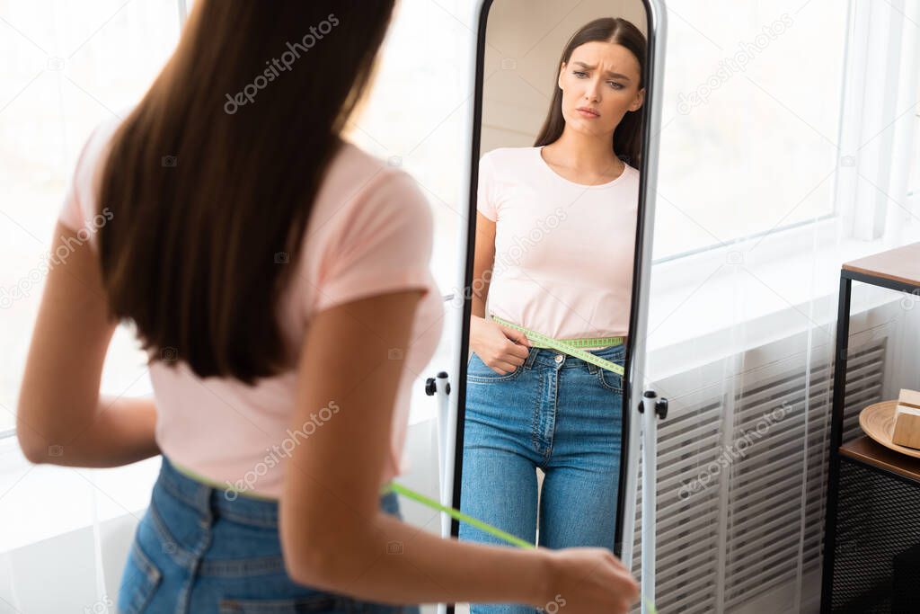 Sad Woman Measuring Waist After Weight Gain Standing At Home