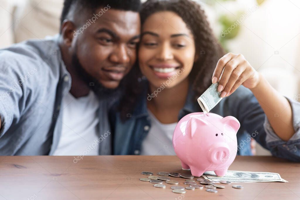 Family Budget. Happy Black Couple Putting Money To Piggybank At Home