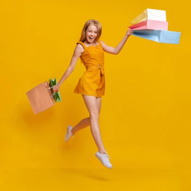 Alışveriş torbaları taşıyan neşeli genç kız, sarı arka planda havaya atlıyor.