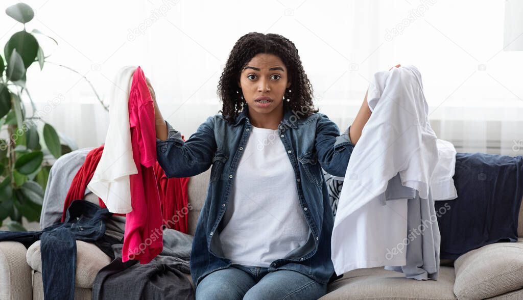 Shocked black girl sitting at home surrounded by lots of clothes