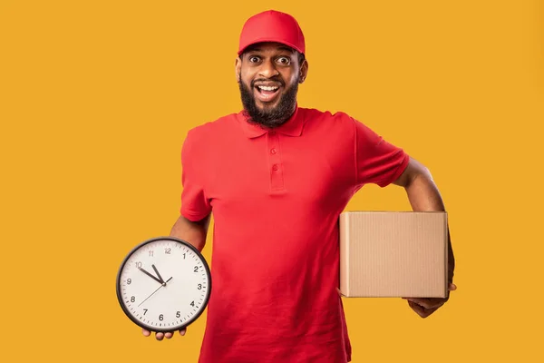 Courier alegre segurando caixa de papelão e relógio sobre fundo amarelo — Fotografia de Stock