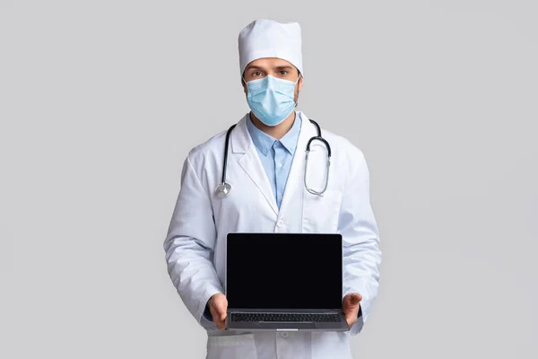 Γιατρός με προστατευτική μάσκα που δείχνει το laptop με κενή οθόνη — Φωτογραφία Αρχείου