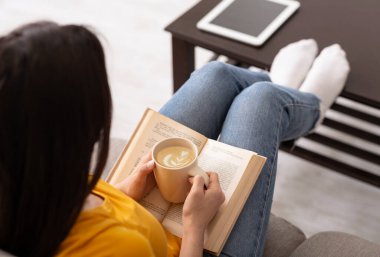 Tanımlanamayan bir kız elinde bir fincan taze aromatik kahve tutuyor ve evde ilginç bir kitap okuyor.