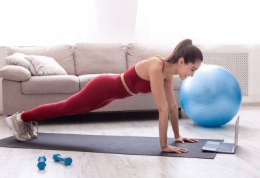 Evde dayanıklılık eğitimi. Kalas egzersizi yapan ya da apartmandaki video sınıfına şınav çeken bin yıllık bir kadın.