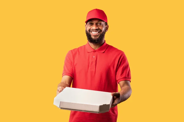 Mensajero africano entregando caja de pizza de pie en el estudio, fondo amarillo — Foto de Stock