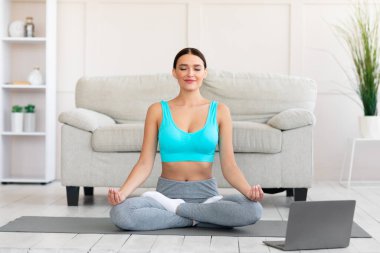 Evde Lotus pozisyonunda oturan kız dizüstü bilgisayarda meditasyon yapıyor.