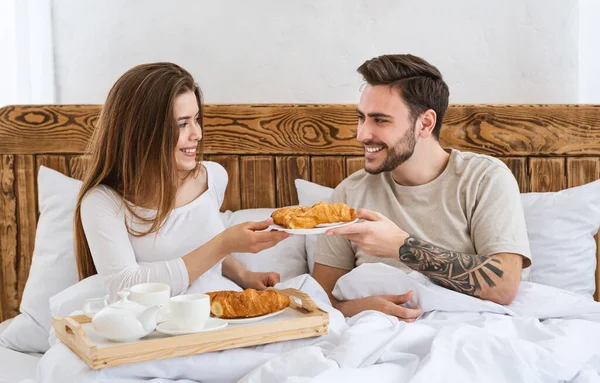 Romantikus reggeli két fogalom. A lány croissant-t ad a férfinak, az ágyban — Stock Fotó