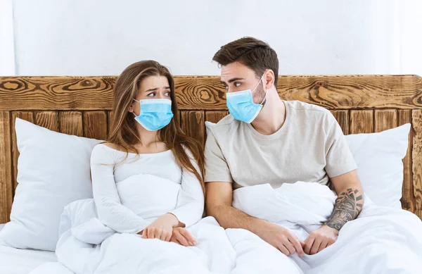 Ασθένεια και καραντίνα. Λυπηρό αγόρι και κορίτσι με προστατευτικές μάσκες κάθονται στο κρεβάτι. — Φωτογραφία Αρχείου