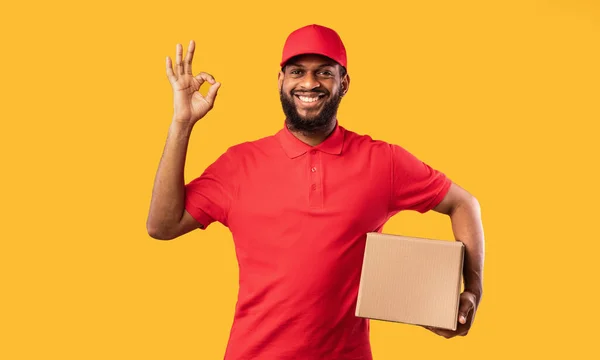 Koerier Guy Holding Kartonnen doos Gesturing OK op gele achtergrond — Stockfoto