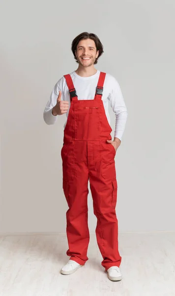 Smiling young laborer posing in work overalls and showing thumb up