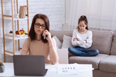 İş annesi dizüstü bilgisayar ve akıllı telefonla çalışırken kızı evde kanepede oturuyor.