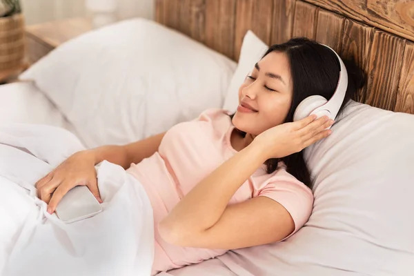 Asiática chica en auriculares escuchar música en cama en casa —  Fotos de Stock