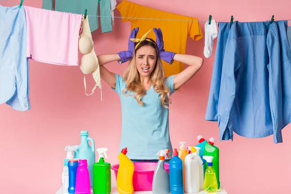 Triste ama de casa sostiene la cabeza con las manos, delante de ella está el lavabo y los suministros de limpieza, detrás está la ropa limpia en la cuerda — Foto de Stock