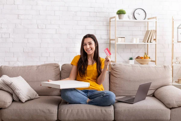 Snelle levering aan huis. Millennial vrouw met creditcard, laptop en afhaalpizza zittend op comfortabele bank binnen — Stockfoto