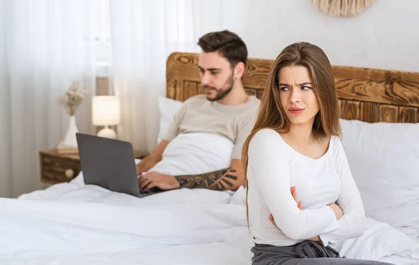 Mężczyzna pracuje na laptopie, kobieta jest obrażona przez łóżko w sypialni. — Zdjęcie stockowe