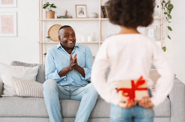 Quédate en casa y celebra. Niña afroamericana sosteniendo presente para el abuelo detrás de su espalda en el interior — Foto de Stock