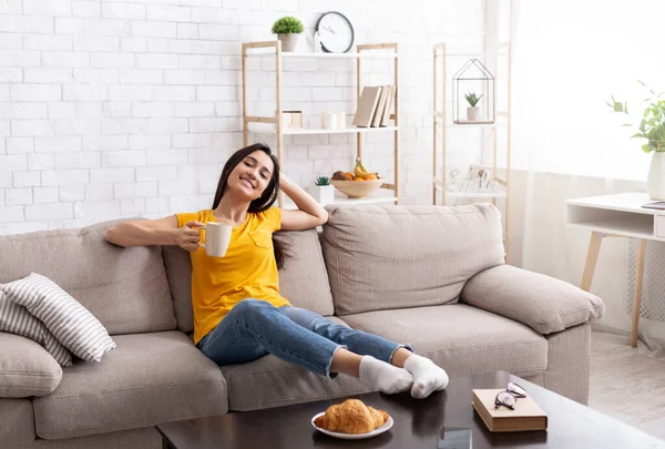 Relaxado menina milenar sentado no sofá com xícara de café em casa, desfrutando de manhã pacífica — Fotografia de Stock