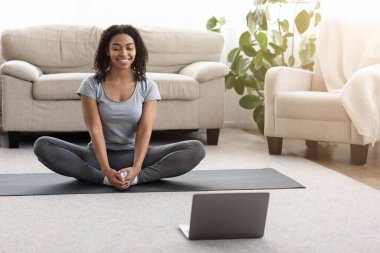 Ev Yogası. Gülümseyen Afrikalı Kadın laptopun önünde evinde meditasyon yapıyor.