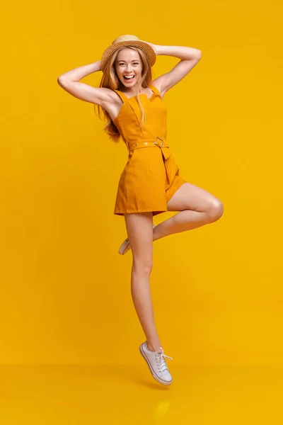 Moda de verano. Chica alegre en mono y sombrero saltando sobre fondo amarillo — Foto de Stock