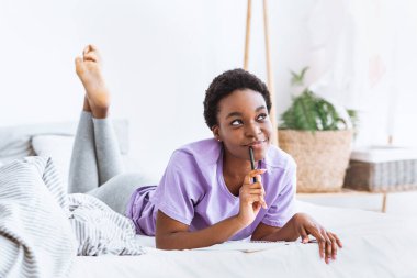 Düşünceli Afrikalı Amerikalı kadın yatağa uzanıyor ve not defterine notlar yazıyor.