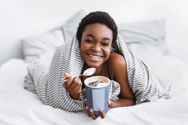 Afroamerykanka leżąca na łóżku, owinięta w koc i jedząca lody — Zdjęcie stockowe