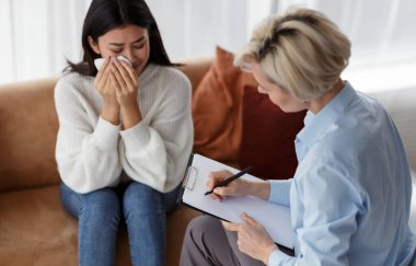 Psikoterapist not alıyor ofiste oturan ağlayan kadını dinliyor.