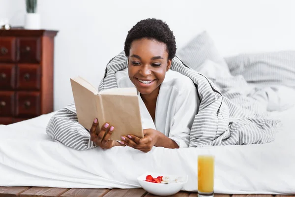 Feliz afroamericana chica leyendo libro en la cama, desayuno cerca —  Fotos de Stock