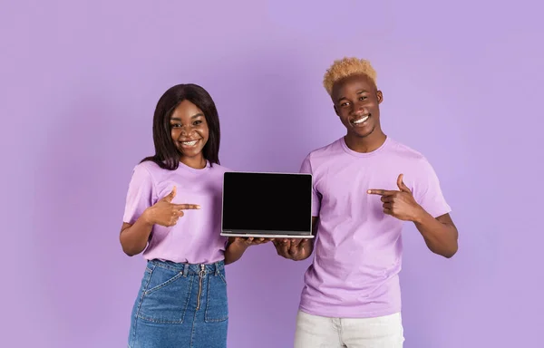 Millennial chica afroamericana y chico apuntando a la pantalla vacía del ordenador portátil en fondo lila, espacio para el diseño del sitio web —  Fotos de Stock