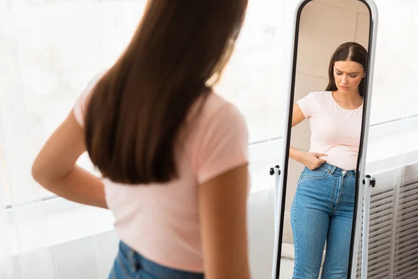 Mulher infeliz ganhando peso tocando barriga em pé no espelho interior — Fotografia de Stock