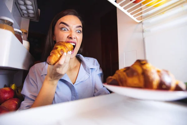 Hladová žena jí croissant z lednice, pohled zevnitř — Stock fotografie