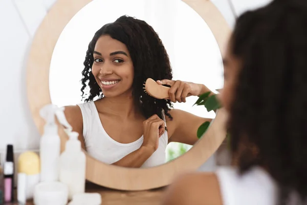 Smiling black woman combing her beautiful curly hair near mirror in bathroom — Stock Photo, Image