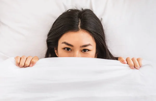 Азиатка выглядывает из одеяла, просыпаясь в постели — стоковое фото