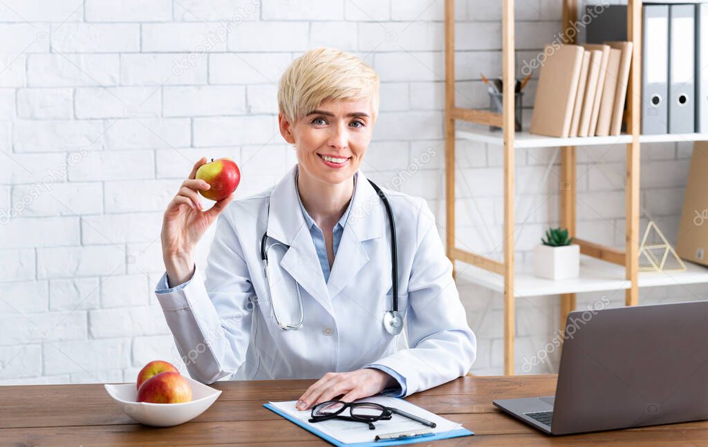 Smiling woman in white coat holds red apple, sitting at table in interior