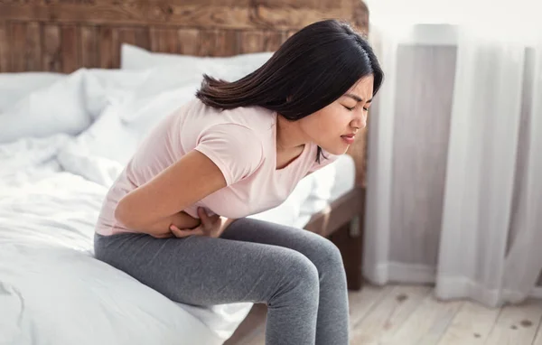 Asiática chica having abdominal dolor sentado en cama en casa —  Fotos de Stock