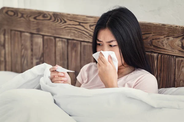 Donna che tiene il termometro che soffia il naso nel tessuto seduto a letto — Foto Stock