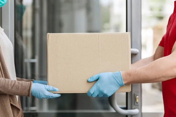 Obsługa klienta podczas kwarantanny. Kurier w rękawiczkach medycznych daje pudełko klientowi przy drzwiach — Zdjęcie stockowe