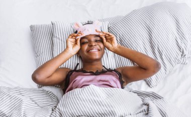 Hafta sonu yatakta. Gülümseyen Afrikalı Amerikalı kız uyku maskesini çıkarıyor.