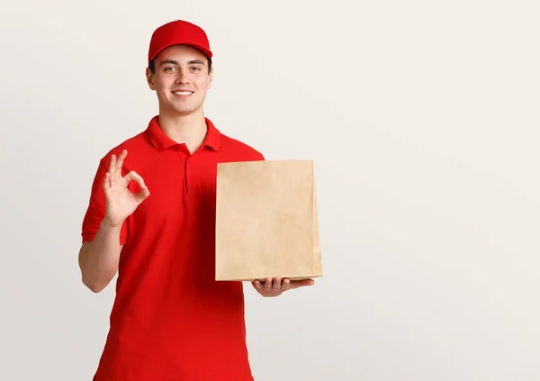 Futár egyenruhában tartja papír csomagot, és azt mutatja, szimbólum rendben kézzel — Stock Fotó