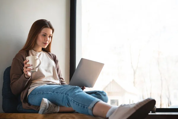 Mujer joven que trabaja en el ordenador portátil mientras está sentado en el alféizar de la ventana —  Fotos de Stock