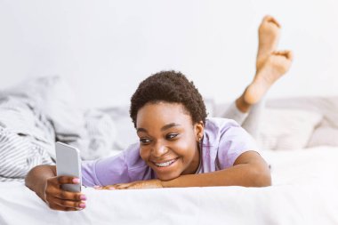 Gülümseyen Afrikalı Amerikalı kız akıllı telefondan bakıyor ve daktilo yazıyor.