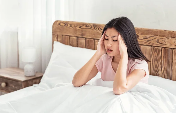 Mulher chinesa com dor de cabeça sofrendo de dor sentada na cama — Fotografia de Stock