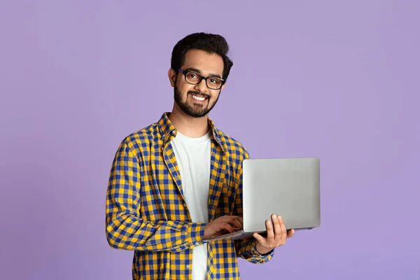 Sonriente chico indio con computadora portátil trabajando o estudiando en línea sobre fondo lila —  Fotos de Stock