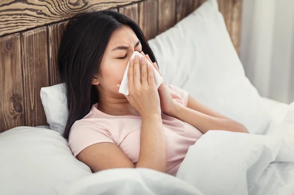 Sjuk kinesisk kvinna blåser näsan i vävnaden ligger i sängen — Stockfoto