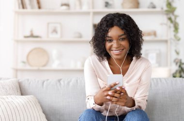 Akıllı telefonlu ve kulaklıklı gülümseyen afro kız evde müzik dinliyor.