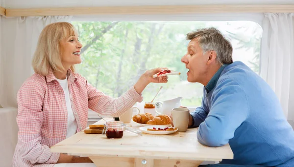 Gemeinsam campen. Schöne reife Frau füttert ihren Mann im Wohnmobil mit Toast und Marmelade — Stockfoto